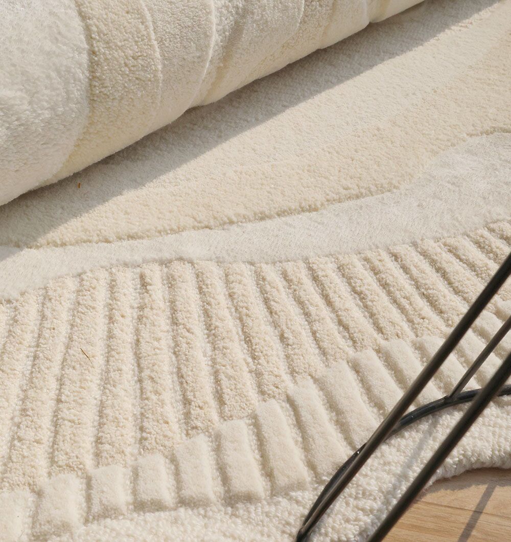A close-up of the RAMSHA HOME Natural Handmade Area Rug - HTWV-126, cream-colored with varied patterns, is partially rolled on a light wood floor. Black metal chair legs appear in the lower right corner.