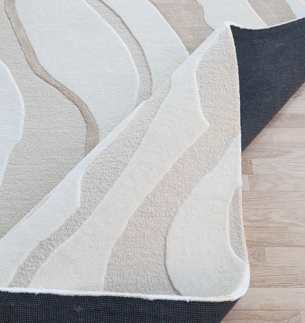 A close-up of the RAMSHA HOME Handcrafted Living Room Area Rug – HTWV-127, featuring abstract wavy patterns in neutral tones, partially lifted to show a black non-slip rug pad underneath on a light wood floor.