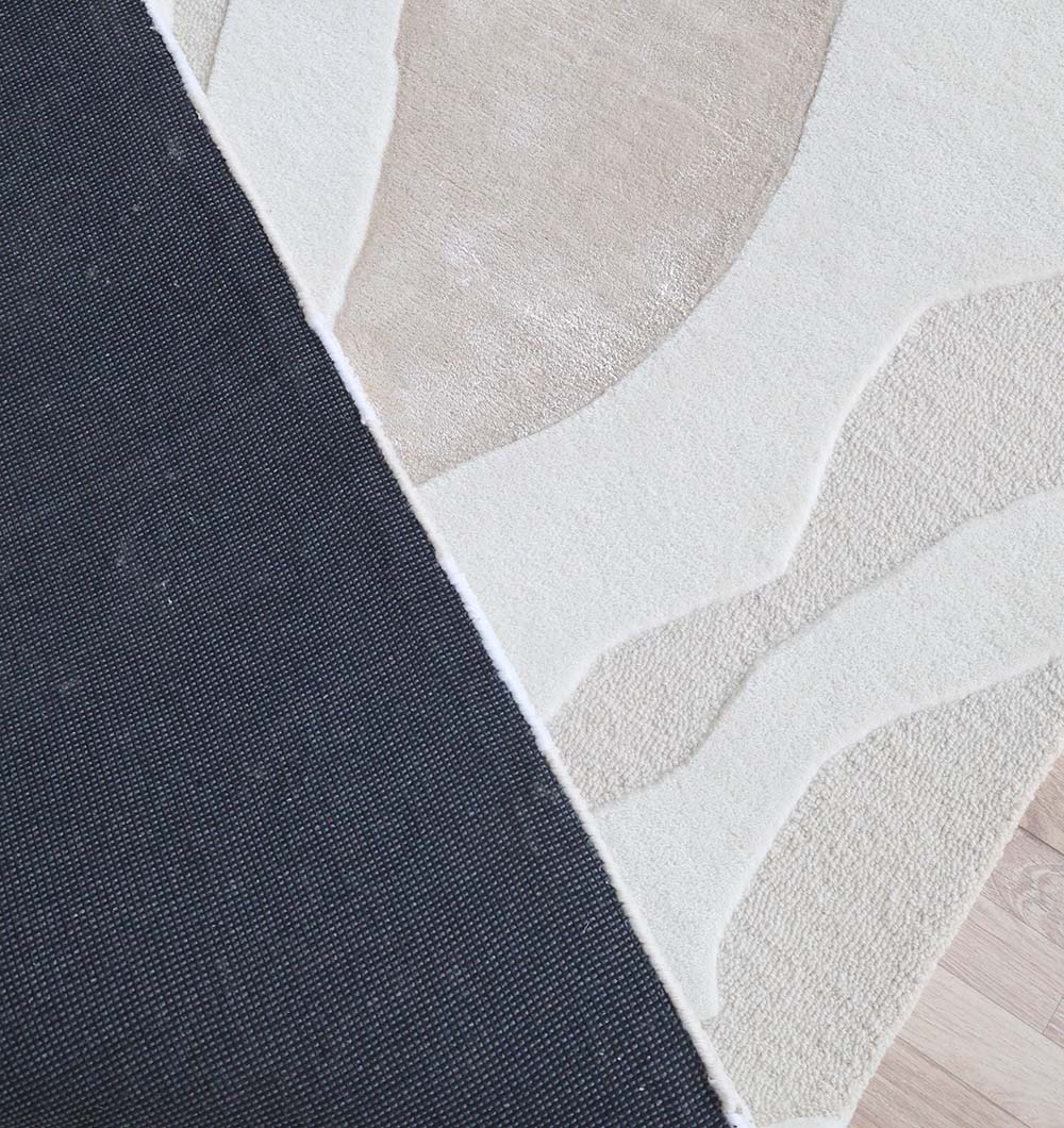 A close-up of the RAMSHA HOME Handcrafted Living Room Area Rug – HTWV-127 shows its textured cream and beige pattern with a dark gray non-slip backing, partially folded on a light wooden floor.