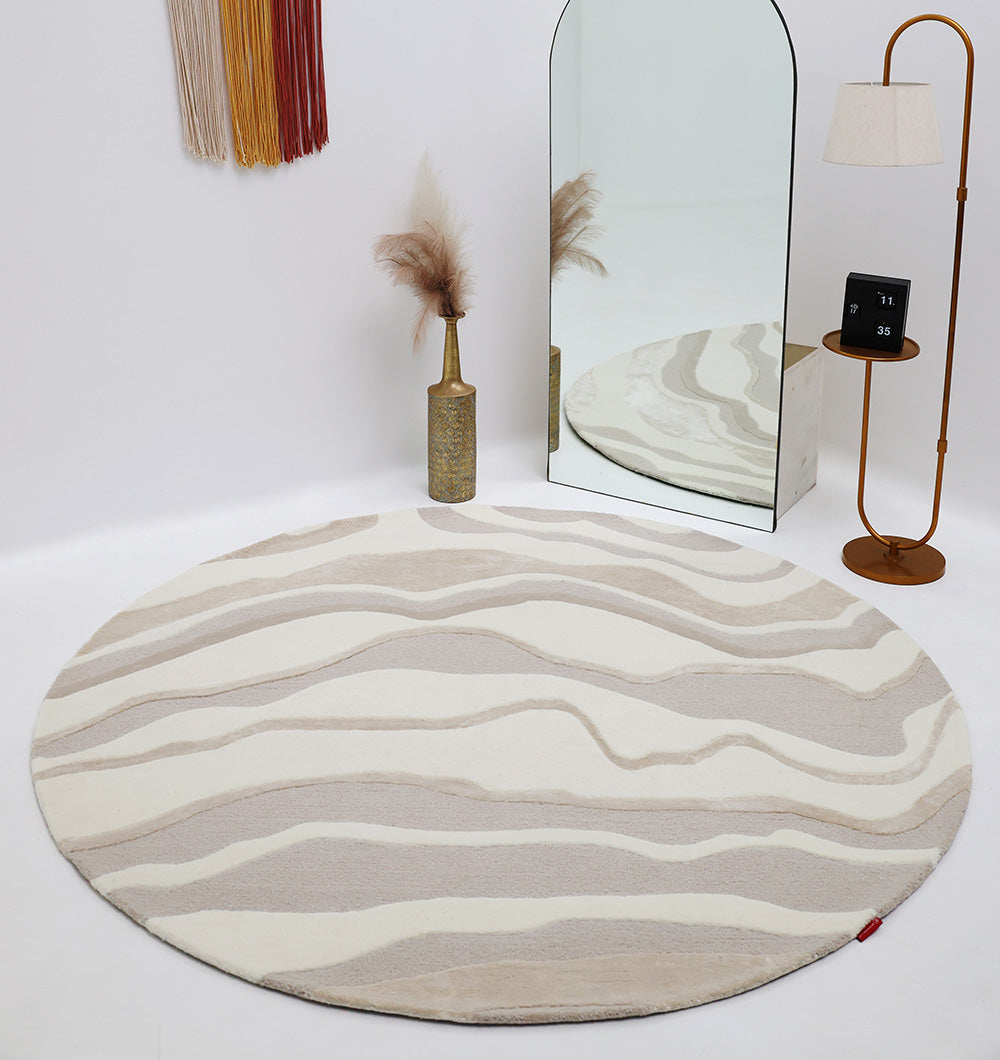 A modern, minimalist room featuring the RAMSHA HOME Handcrafted Living Room Area Rug – HTWV-127 in beige and white wavy patterns, complemented by a tall mirror, standing lamp, gold vase with pampas grass, and earth-toned wall textile art.