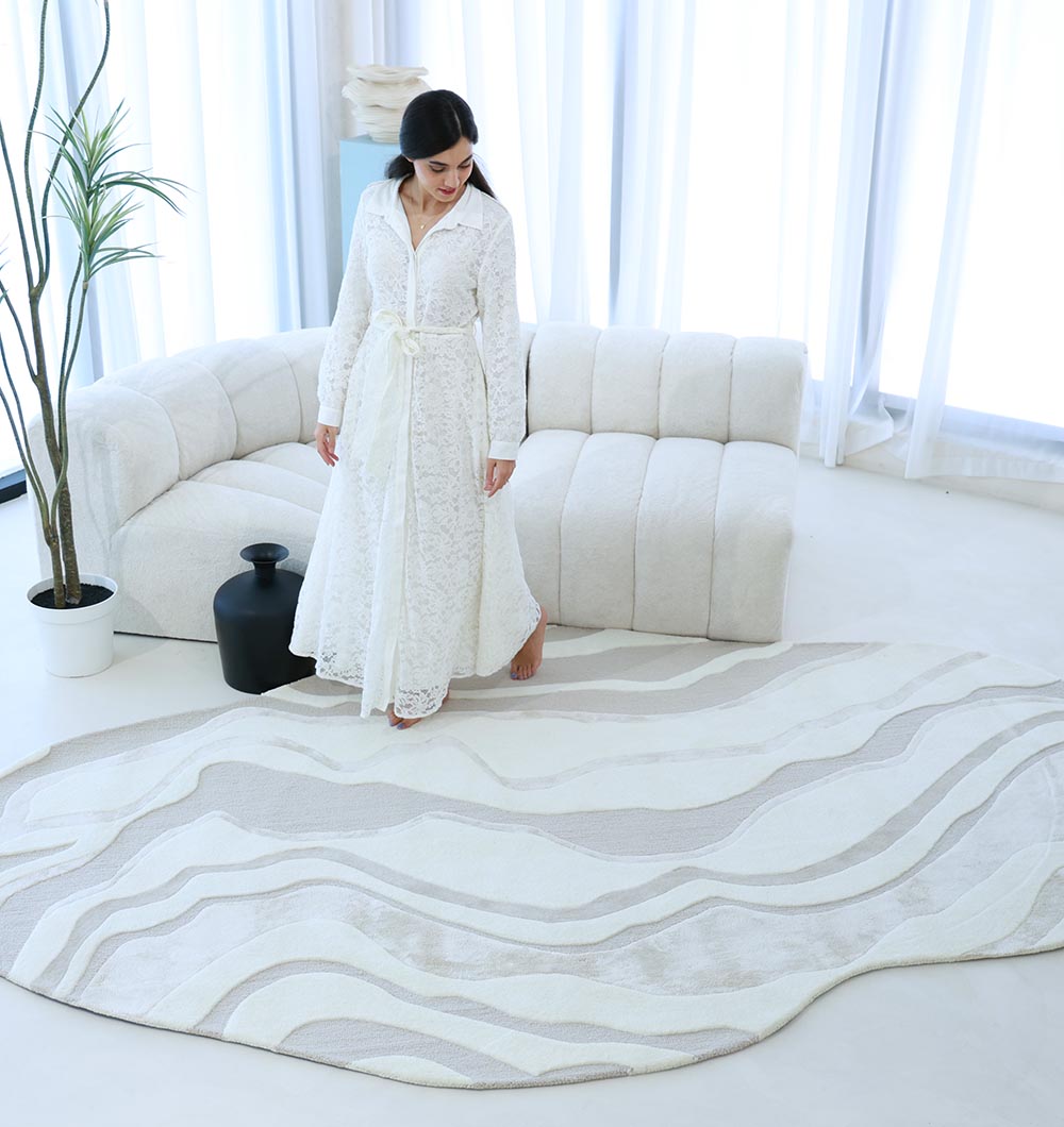 A woman in a long white lace dress stands barefoot on the RAMSHA HOME Handcrafted Living Room Area Rug – HTWV-127 in a bright, modern living room featuring a curved cream sofa, black vase, potted plant, and large windows with sheer curtains.