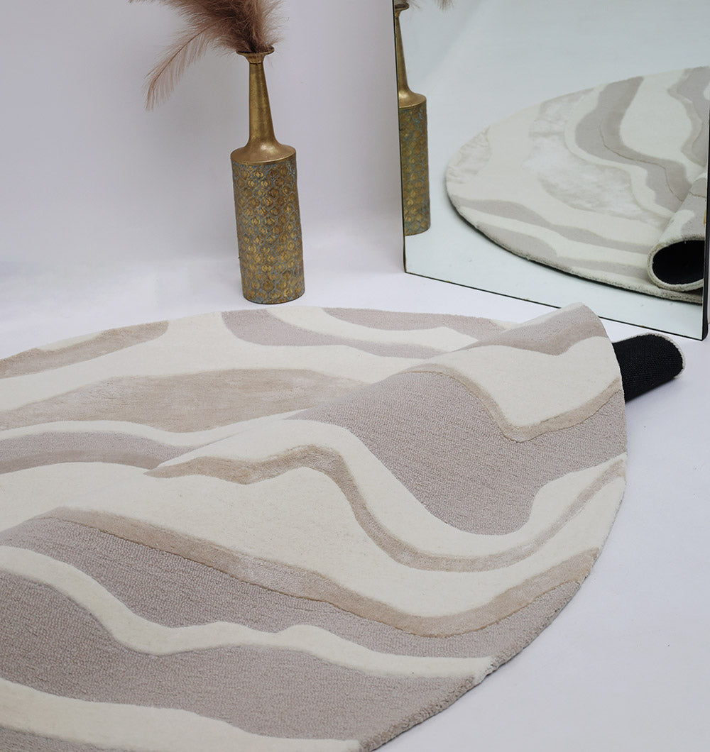 The RAMSHA HOME Handcrafted Living Room Area Rug – HTWV-127, a modern abstract piece in neutral tones, is partially rolled next to a gold vase with dried pampas grass and a mirror reflecting both the rug and vase in the background.