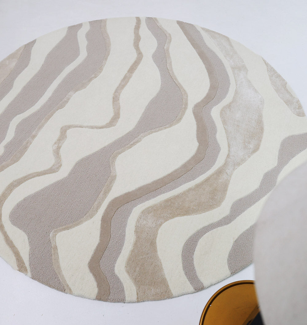 The RAMSHA HOME Handcrafted Living Room Area Rug – HTWV-127, featuring wavy beige and cream patterns, adds elegance to a white floor. A hint of a golden object is visible in the lower right corner.