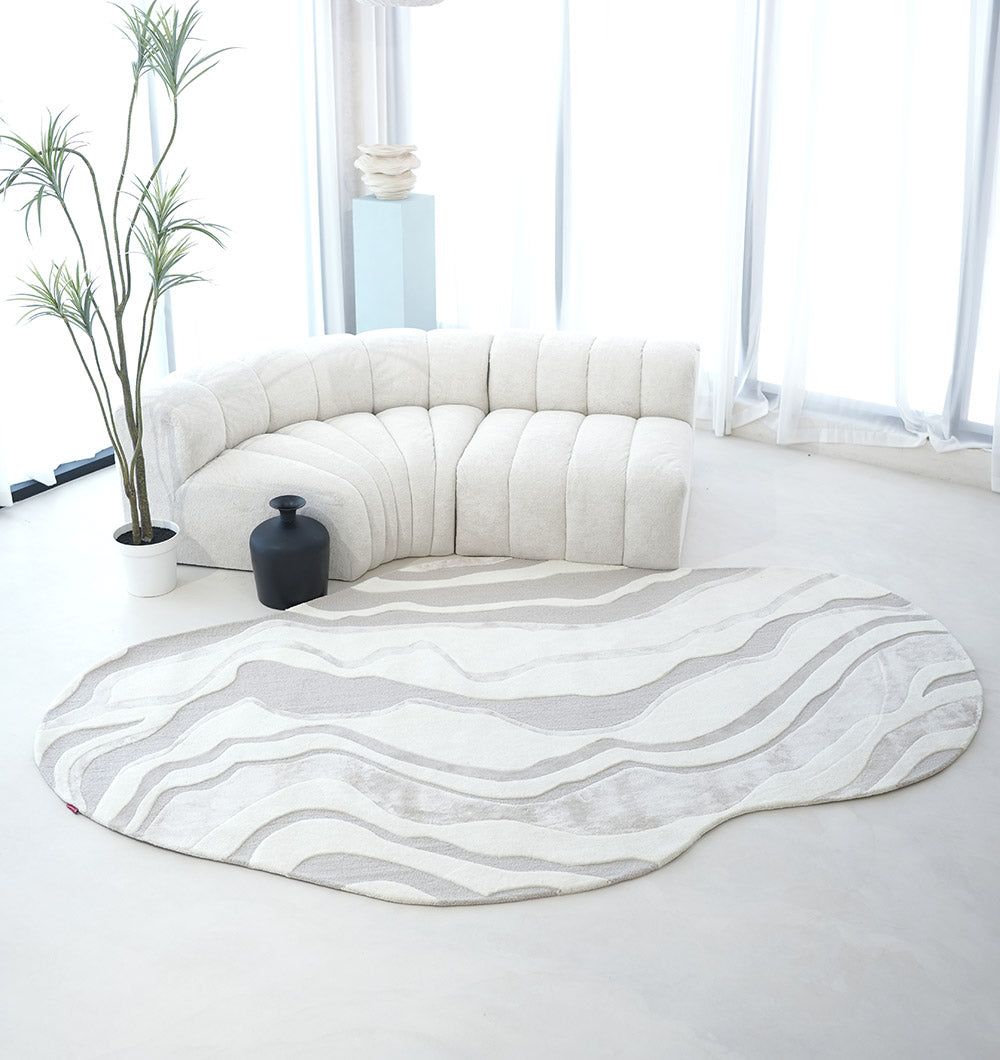 A modern, minimalist living room featuring a curved white tufted sofa, RAMSHA HOME's Handcrafted Living Room Area Rug – HTWV-127 with wavy beige and white patterns, a potted plant, a black vase, and large windows with sheer white curtains.