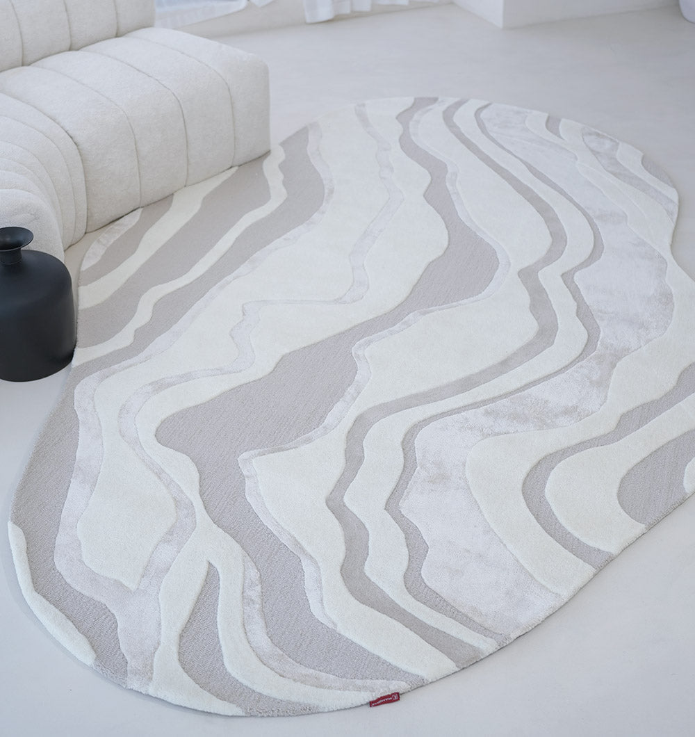 The RAMSHA HOME Handcrafted Living Room Area Rug – HTWV-127, featuring modern abstract wavy cream and light gray patterns, is placed on a light floor beside a curved cream sofa and a small black side table.
