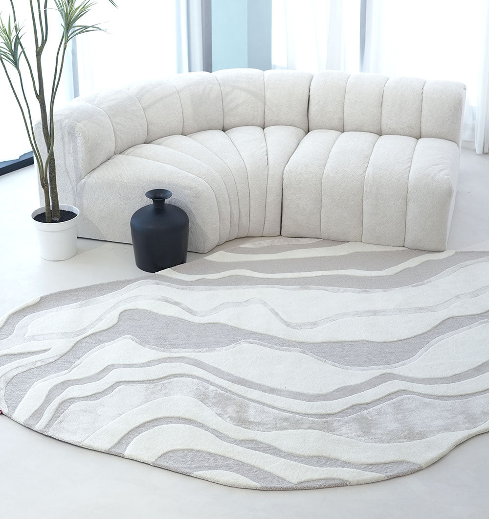 A modern living room features a white curved sectional, a black floor vase, a tall potted plant, and the RAMSHA HOME Handcrafted Living Room Area Rug – HTWV-127 in beige and white. Sunlight streams in through large windows behind the sofa.