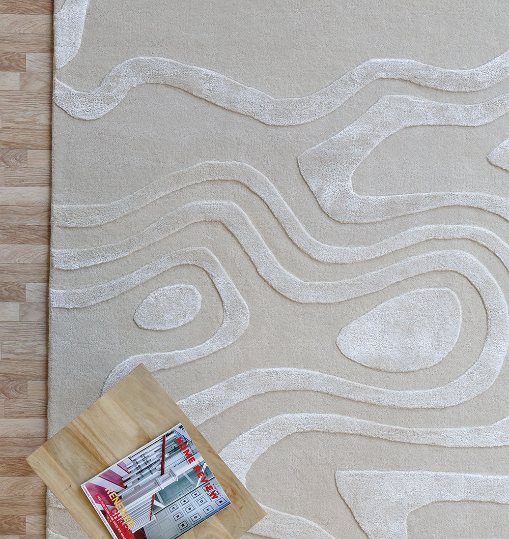 The RAMSHA HOME Topographic Wave Pattern Beige & Ivory Rug – HTWV 128, made from a wool-viscose blend with raised wavy designs, lies on a light wood floor. A small wooden table in the corner displays a magazine and calculator.