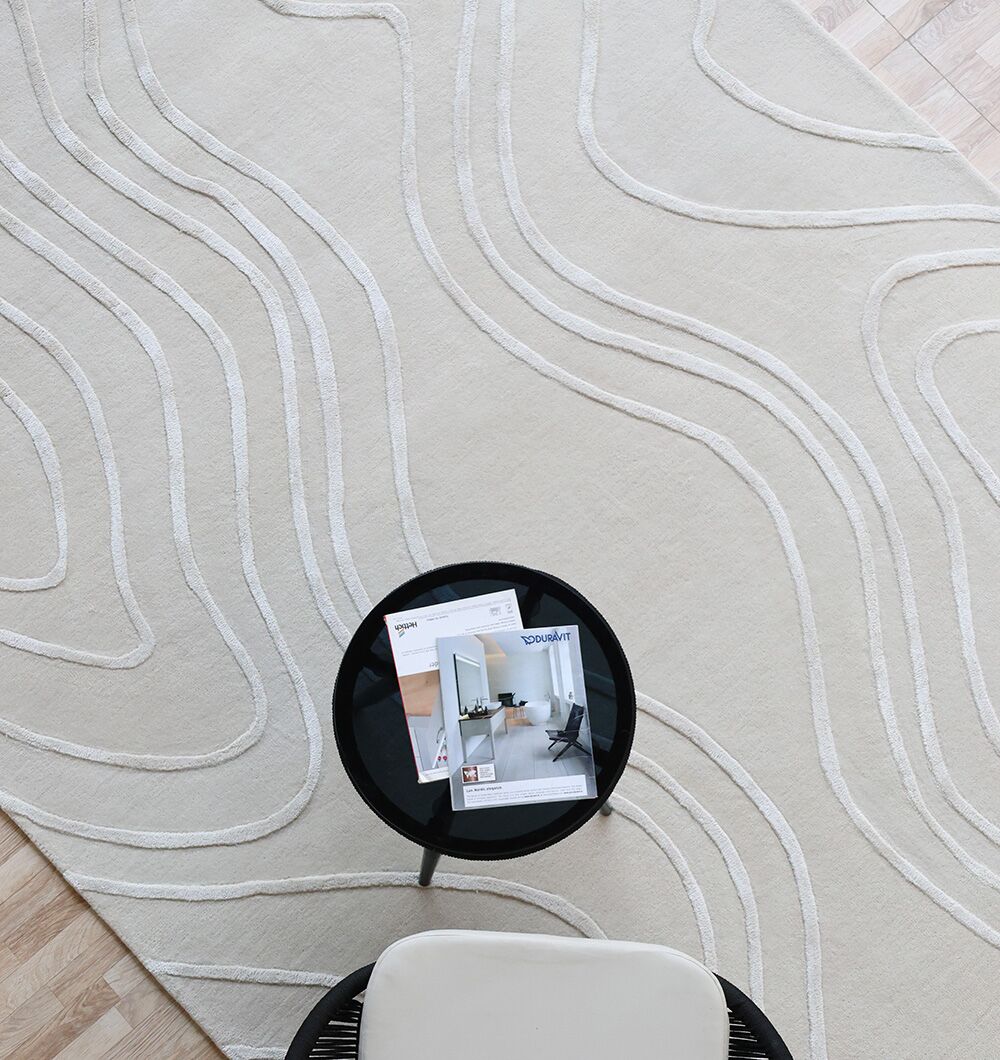 A small black round table with magazines sits on a RAMSHA HOME Multi Color Handmade Area Rug (HTWV-133), featuring abstract wavy lines, next to a white cushioned chair on a light wood floor.