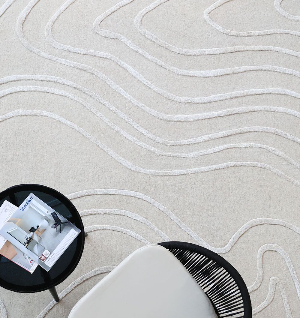 The RAMSHA HOME Multi Color Handmade Area Rug - HTWV-133 in cream with wavy raised lines lies under a small round black table with magazines and part of a light chair featuring black woven backrest details.