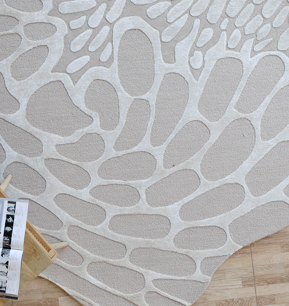 The RAMSHA HOME Organic Flow Hand-Tufted Rug – Soothing Beige Textured Elegance HTWV-134 features an irregular cream and light gray pebble-like pattern, adding subtle texture to wooden floors. A small rack with magazines is partially visible nearby.
