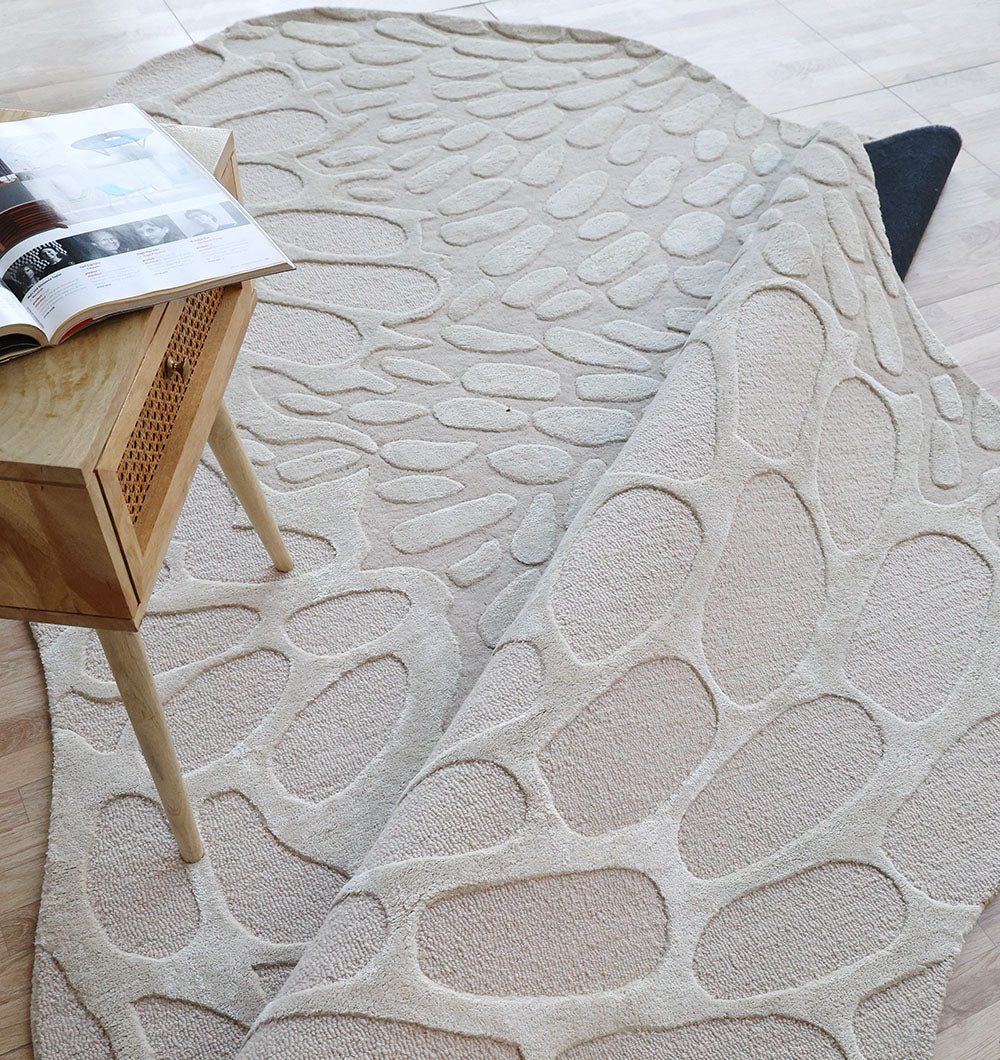 The RAMSHA HOME Organic Flow Hand-Tufted Rug – Soothing Beige Textured Elegance HTWV-134 is partially rolled up on a light wood floor beside a small wooden side table holding an open magazine.
