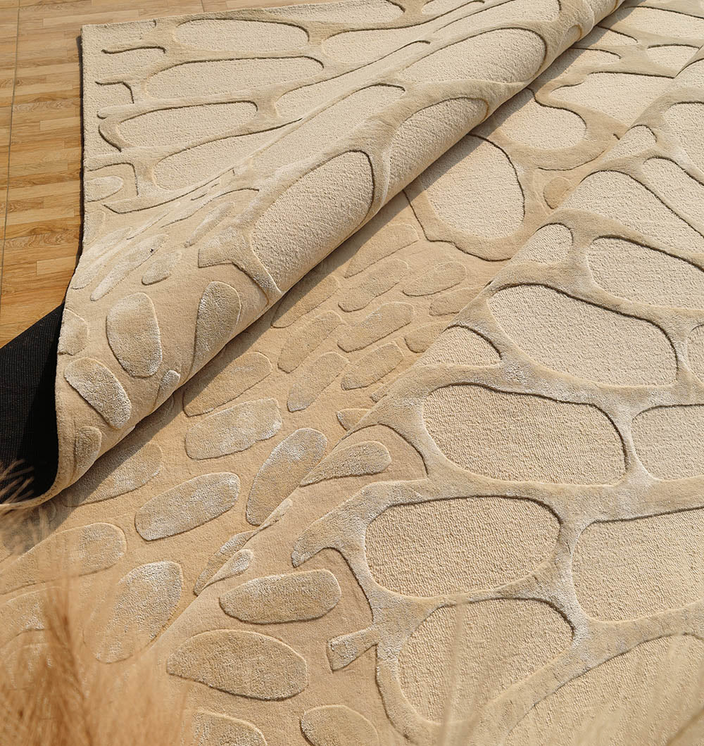 A close-up of the RAMSHA HOME Organic Flow Hand-Tufted Rug – Soothing Beige Textured Elegance HTWV-134, featuring a raised abstract pattern and black backing, partially rolled back on a light wooden floor.