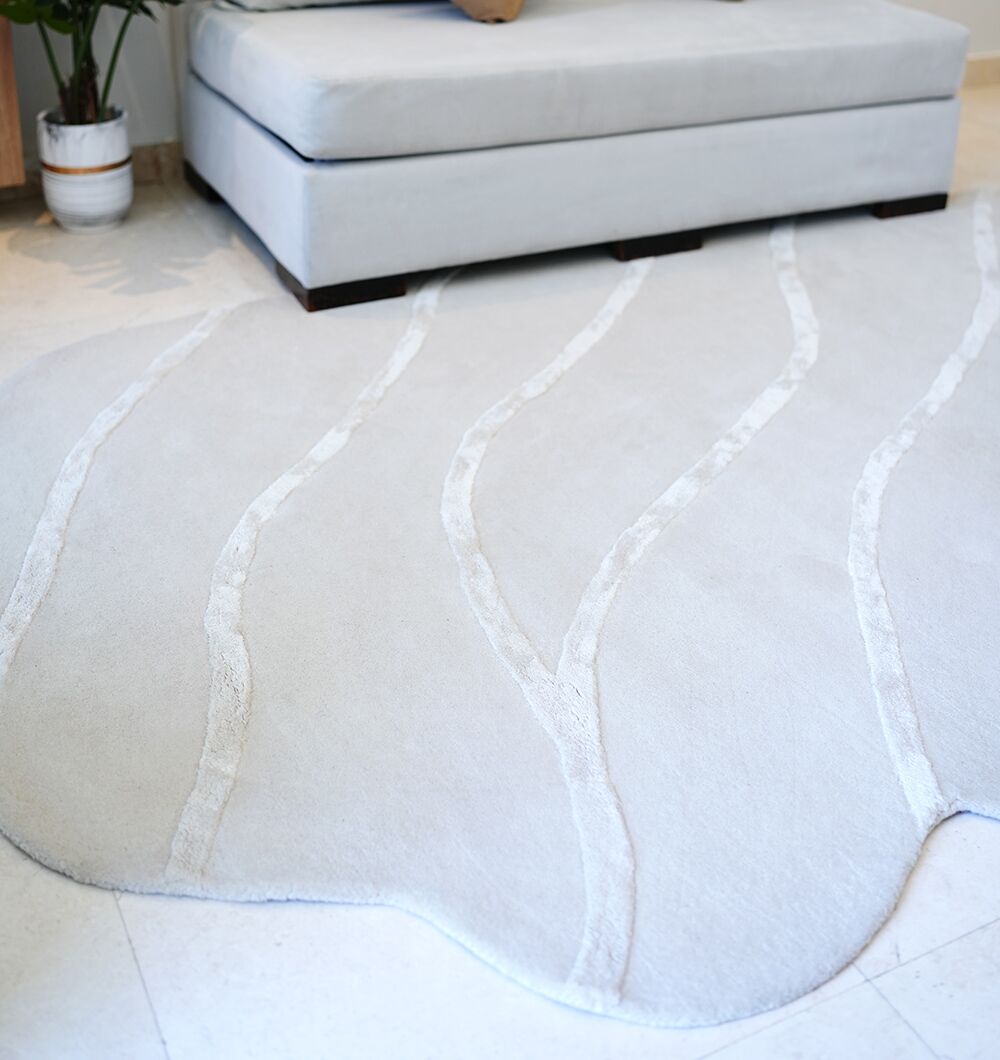 A Multi Color Handmade Area Rug - HTWV-136 (200x300 cm) by RAMSHA HOME with light gray wavy edges and subtle raised lines lies on a cream floor, partly under a modern light gray sofa. A plant sits in the corner.