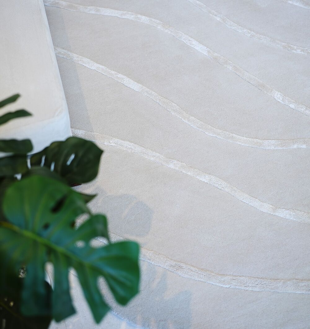 A close-up of the RAMSHA HOME Multi Color Handmade Area Rug (HTWV-136, 200x300 cm) showcases its subtle wavy lines beside a cream sofa, with a green monstera plant in the foreground.