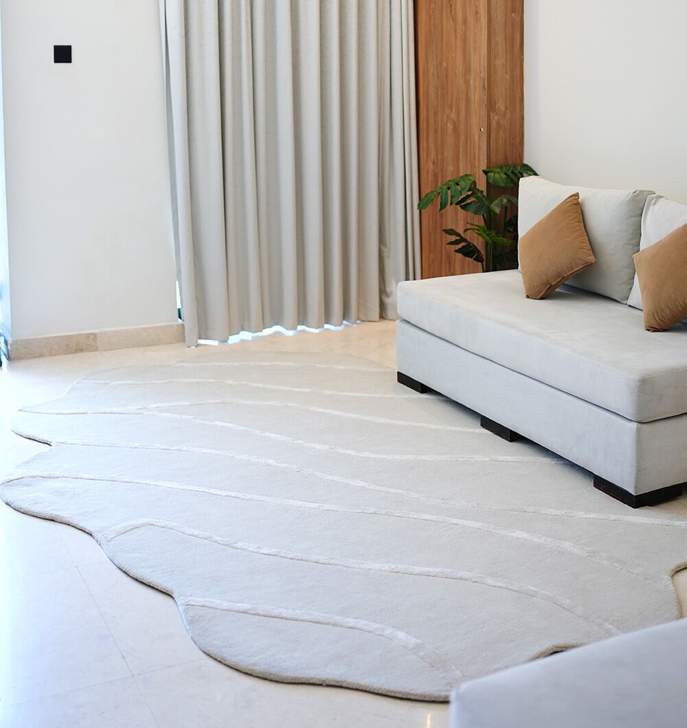 A modern living room with a light grey curved sofa, two brown cushions, the RAMSHA HOME Multi Color Handmade Area Rug - HTWV-136 (200x300 cm), cream floor tiles, beige curtains, a wooden door, and a green plant in the corner.