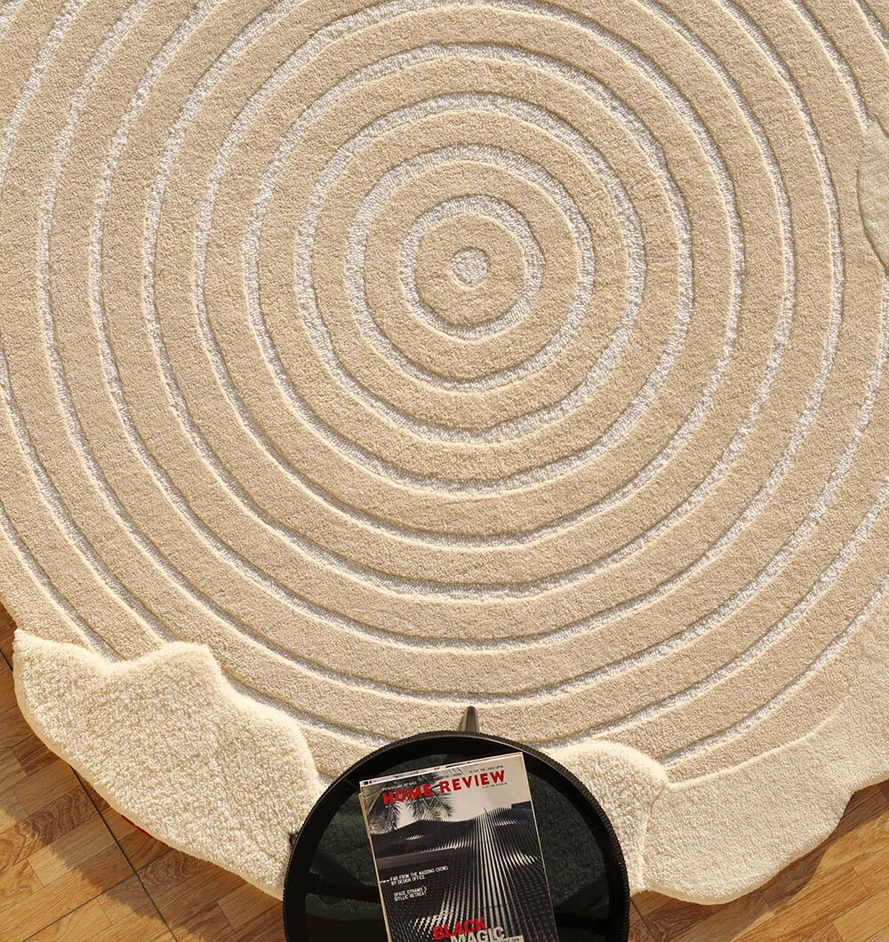 A Natural White Color Handmade Area Rug - HTWV-138 (183 x 200 cm) by RAMSHA HOME with concentric circular patterns lies on a wooden floor. A black side table in front holds a magazine titled Book Review.