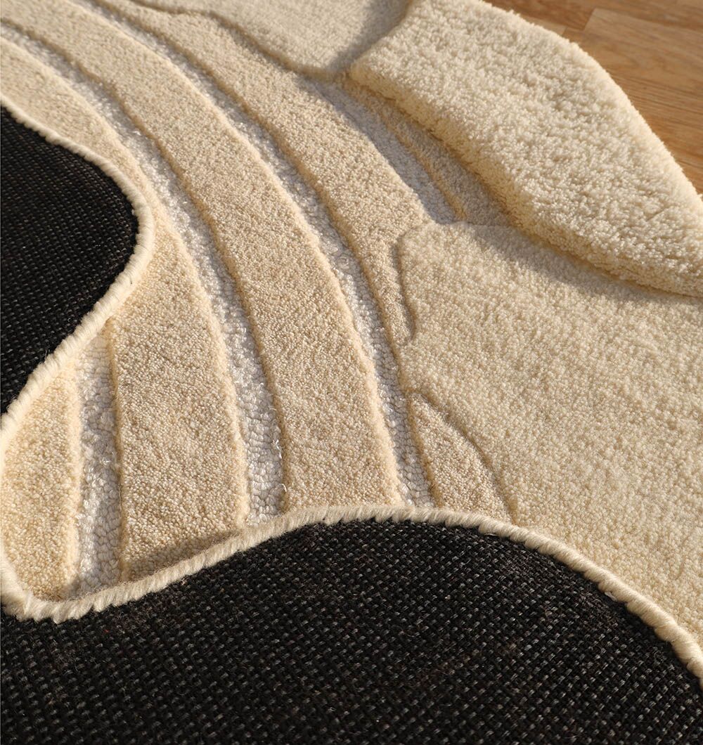 Close-up of the RAMSHA HOME Natural White Color Handmade Area Rug - HTWV-138 (183 x 200 cm) showing its irregular shape, raised cream and beige sections, flat black area, and curved abstract patterns on a wooden floor.