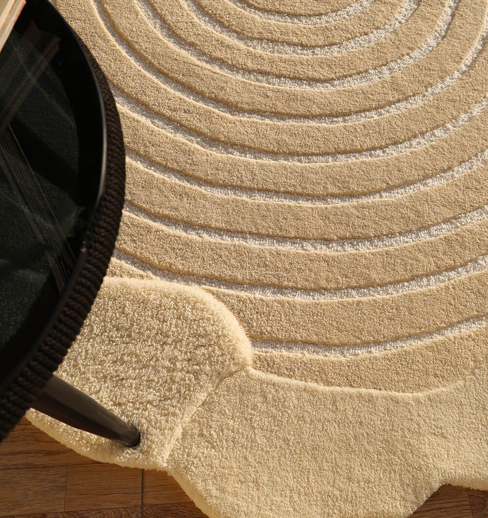 A close-up of the RAMSHA HOME Natural White Color Handmade Area Rug - HTWV-138 (183 x 200 cm) with concentric circles, shown beside a round black table leg on a wooden floor.