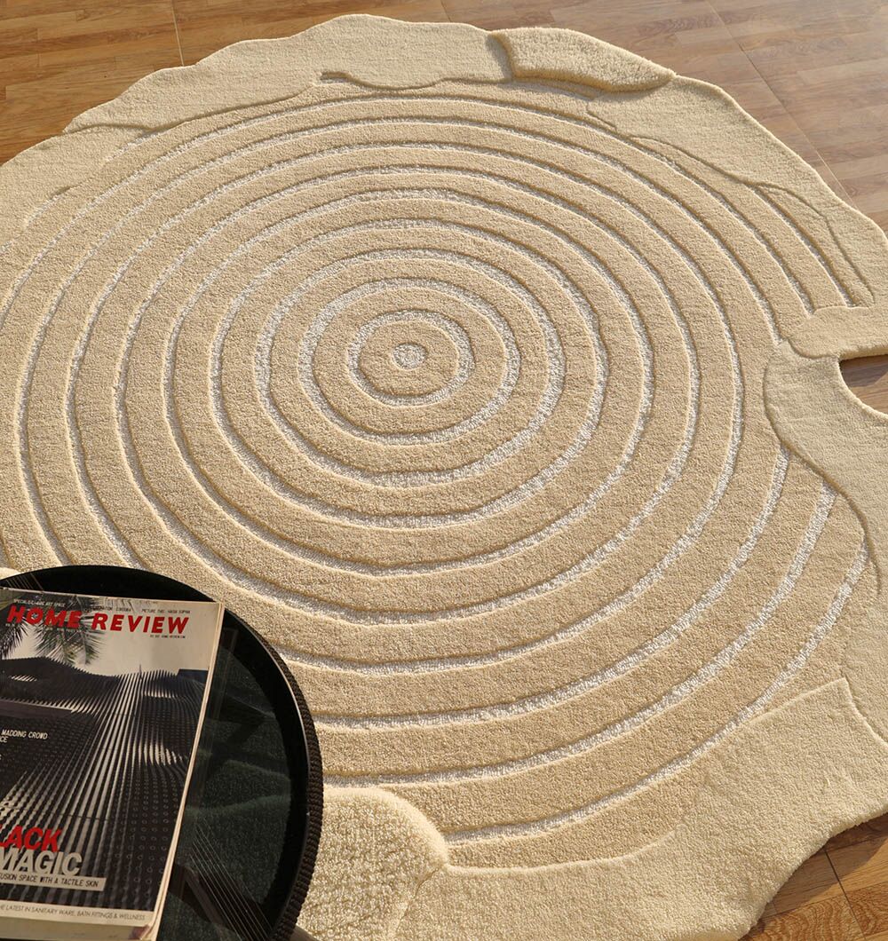 The RAMSHA HOME Natural White Color Handmade Area Rug - HTWV-138 (183 x 200 cm), featuring concentric raised rings like a tree trunk, lies on a wooden floor beside a black round table with a magazine.