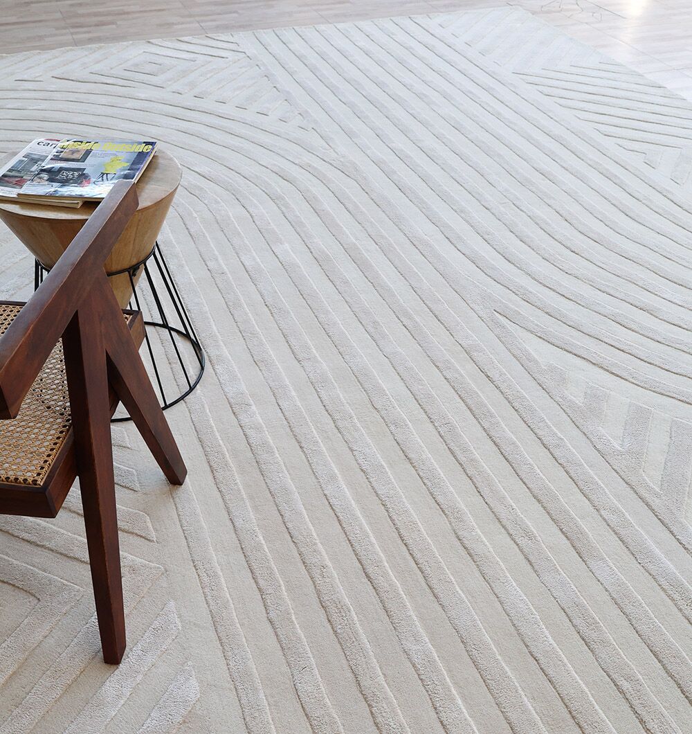 A close-up of the RAMSHA HOME Natural White Color Handmade Area Rug - HTWV-142 (300x400 cm) features geometric patterns. A wooden chair with a woven seat and a small round table with magazines sit atop the ivory rug.