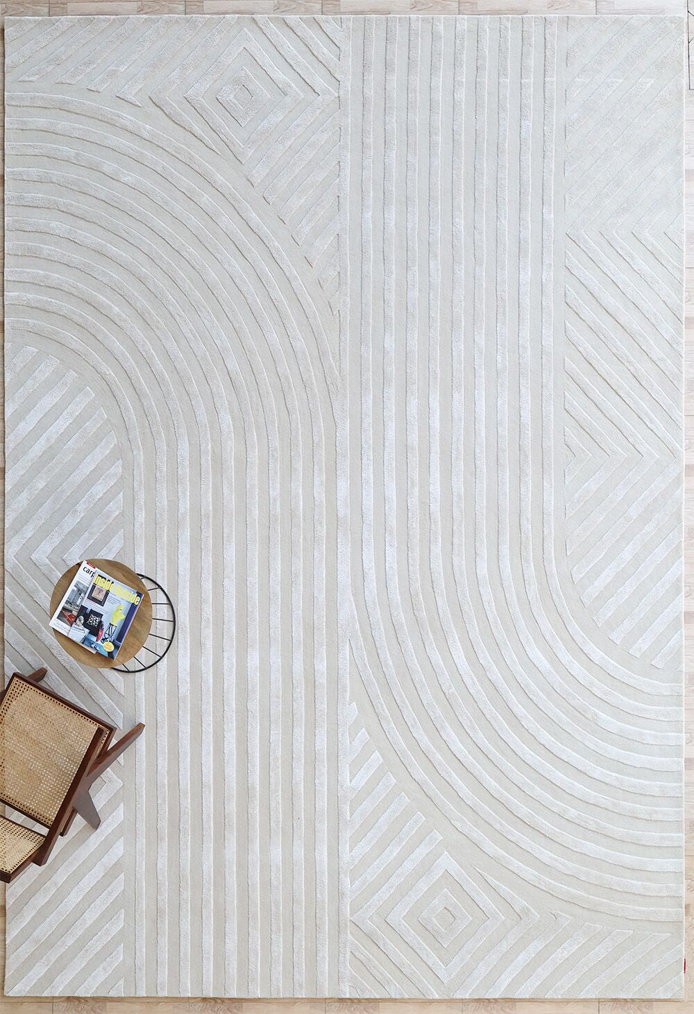 The RAMSHA HOME Natural White Color Handmade Area Rug (HTWV-142, 300x400 cm) brings subtle geometric texture. In the lower left corner, a wooden chair and a small round side table with magazines complete the setting.