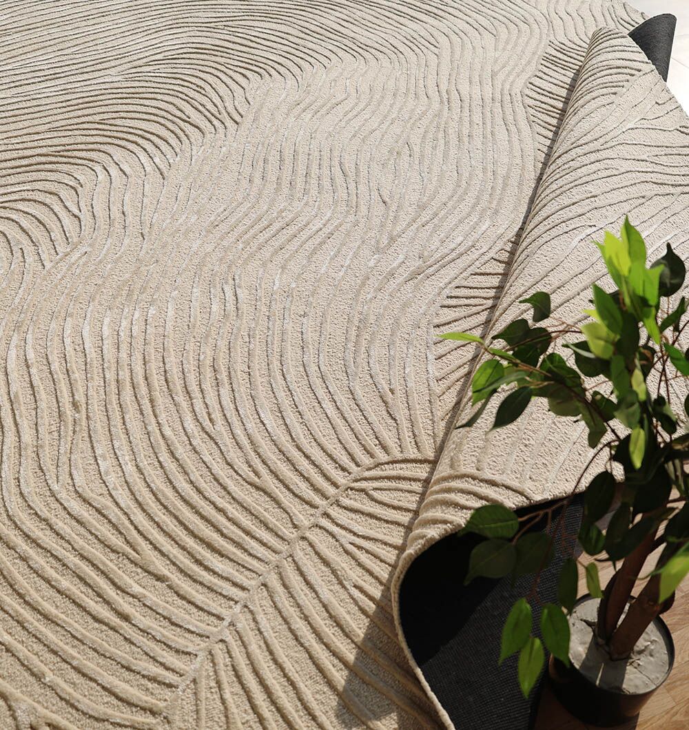 The Natural White Color Handmade Area Rug - HTWV-143 (300x400 cm) by RAMSHA HOME features textured, wavy patterns. Partially folded at the corner, it’s styled with a green potted plant on a wooden floor for a fresh, modern look.