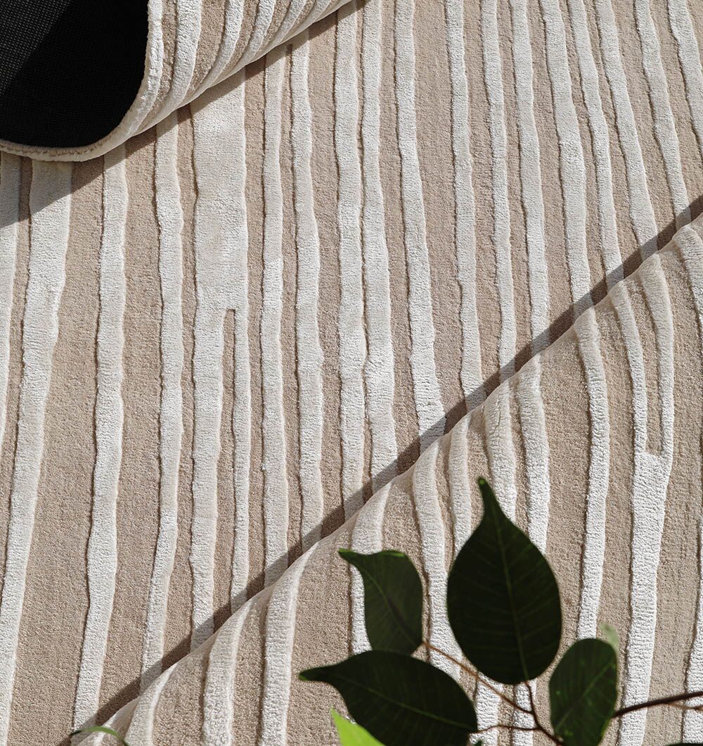 A close-up of the RAMSHA HOME Natural Color Handmade Area Rug - HTWV-147 (300x400 cm), showcasing its beige and white wavy stripes. Partially rolled, it’s complemented by a green leafy plant in the lower right corner for a fresh touch.