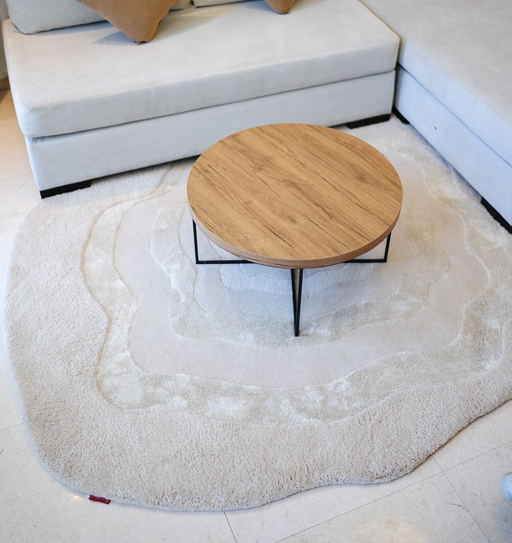 A round wooden coffee table with black legs sits on the RAMSHA HOME Multi Color Handmade Area Rug - HTWV 148, positioned in front of a light-colored sectional sofa in a modern living room.