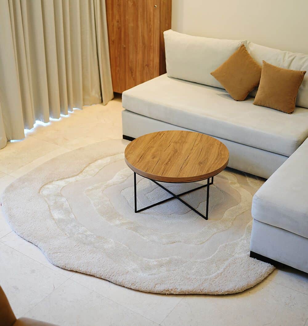 A modern living room featuring a light sectional sofa, two tan cushions, a round wooden coffee table with black legs, and the RAMSHA HOME Multi Color Handmade Area Rug - HTWV 148 on light tile flooring. Curtains cover the window.