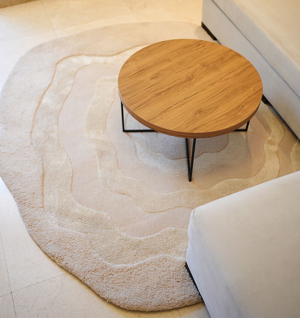 A round wooden coffee table with black metal legs sits on the RAMSHA HOME Multi Color Handmade Area Rug - HTWV 148 next to a white sofa in a bright, modern living room.