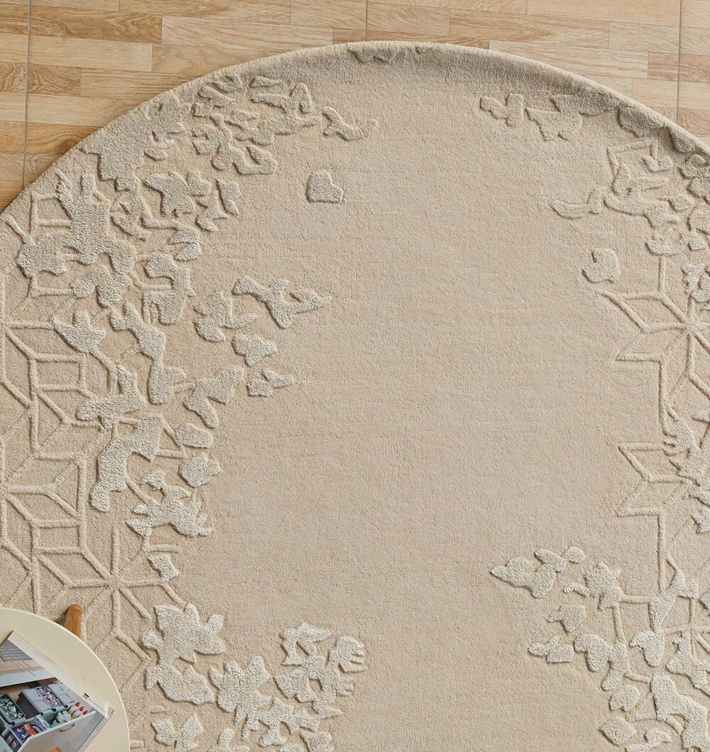 The RAMSHA HOME Natural Color Handmade Rug - HTWV-151 (200 cm), with raised floral and geometric patterns in beige, lies on a light wood floor. A round table with a magazine appears partially in the bottom left corner.