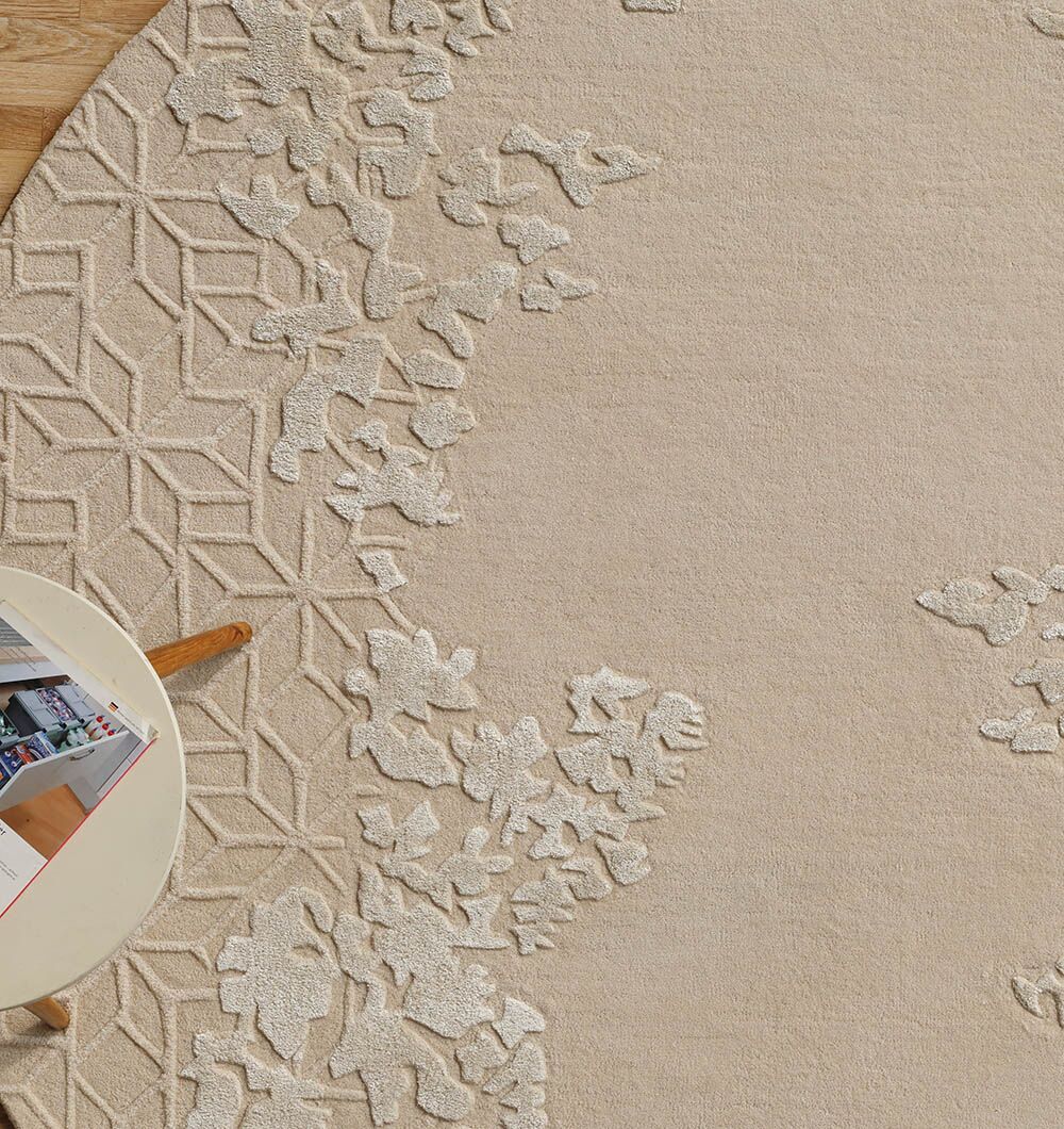 The RAMSHA HOME Natural Color Handmade Rug - HTWV-151 (200 cm) in beige has textured, raised floral and geometric edge patterns. A small white table with an open magazine sits partly on the rug, exposing the wooden floor below.