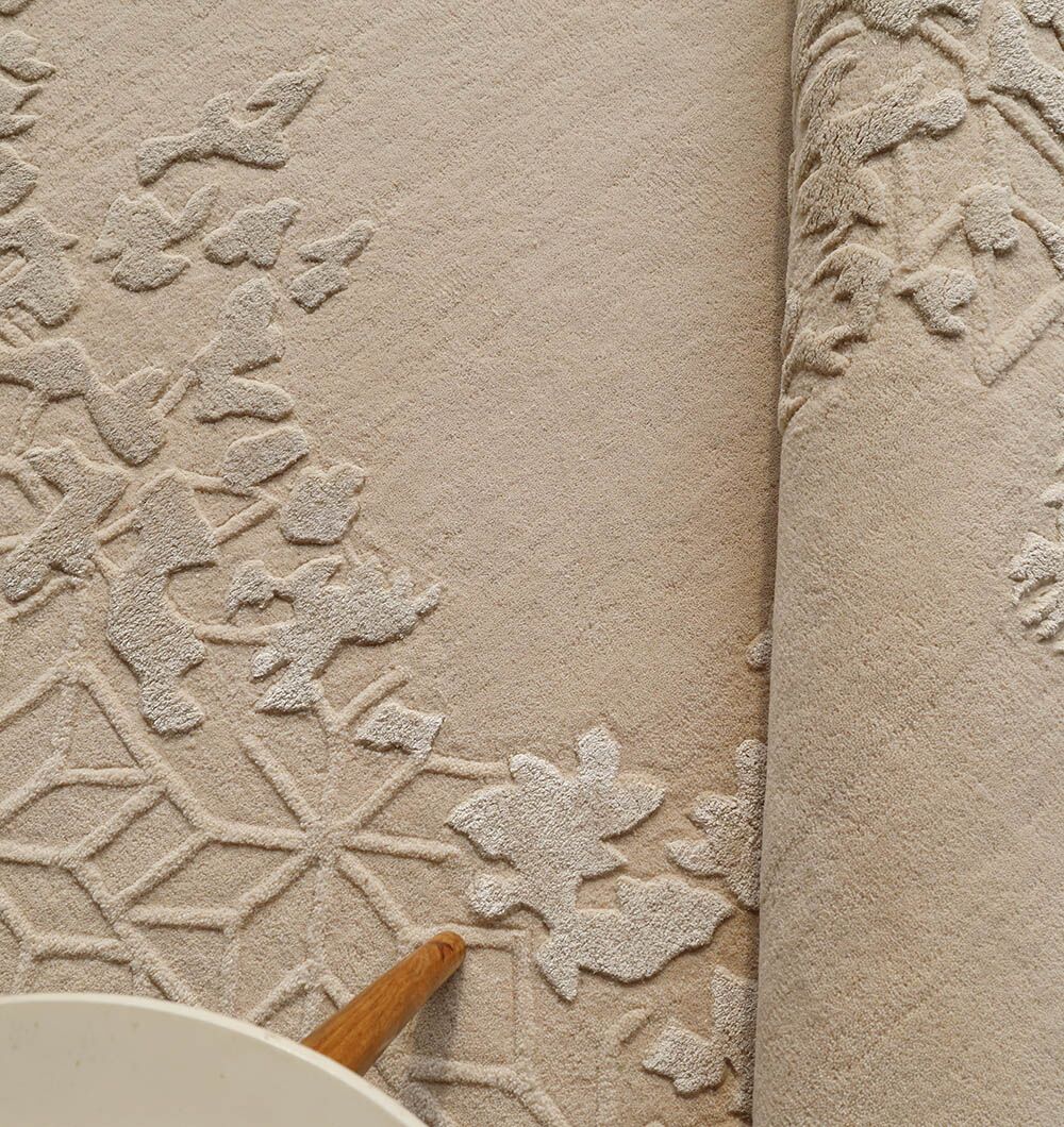 A close-up of the RAMSHA HOME Natural Color Handmade Rug - HTWV-151 (200 cm), featuring carved wool-viscose with raised floral and geometric patterns, is partially rolled up. A light-colored round object, possibly a table, appears in the corner.