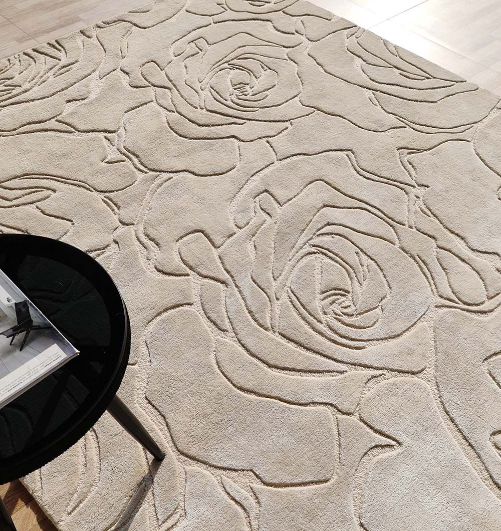 The RAMSHA HOME Home Decor Handmade Area Rug - HTWV-156 features a beige floral design with raised, carved abstract roses. A round black table with magazines sits on its corner, contrasting beautifully against wooden flooring in the background.