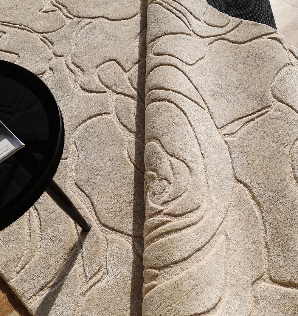 The RAMSHA HOME Home Decor Handmade Area Rug - HTWV-156 features a beige floral design with a carved texture, partially rolled up on a light wooden floor, complemented by a round black table near its elegant raised pattern.