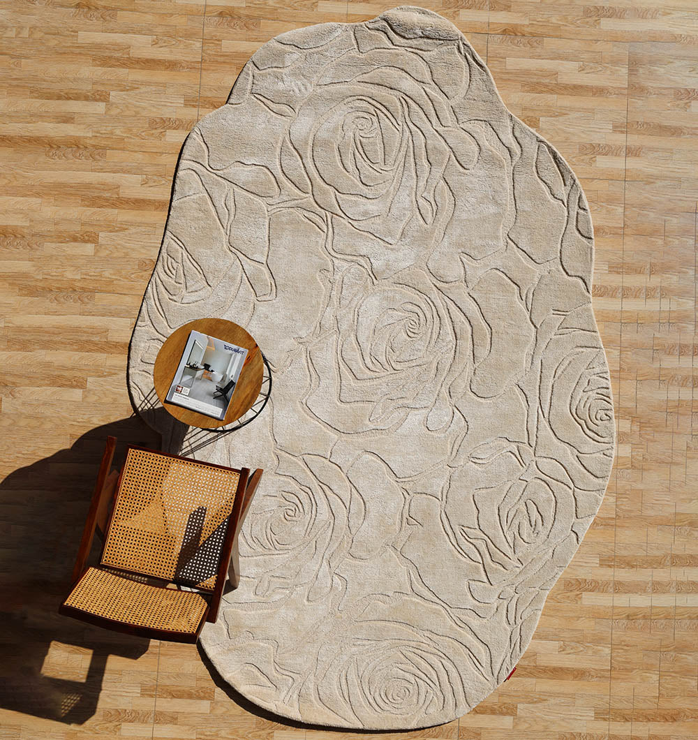 The RAMSHA HOME Handmade Area Rug (HTWV-156), beige with embossed rose patterns and a carved texture, is displayed on a wooden floor with a wooden chair and a small round side table holding an open magazine.