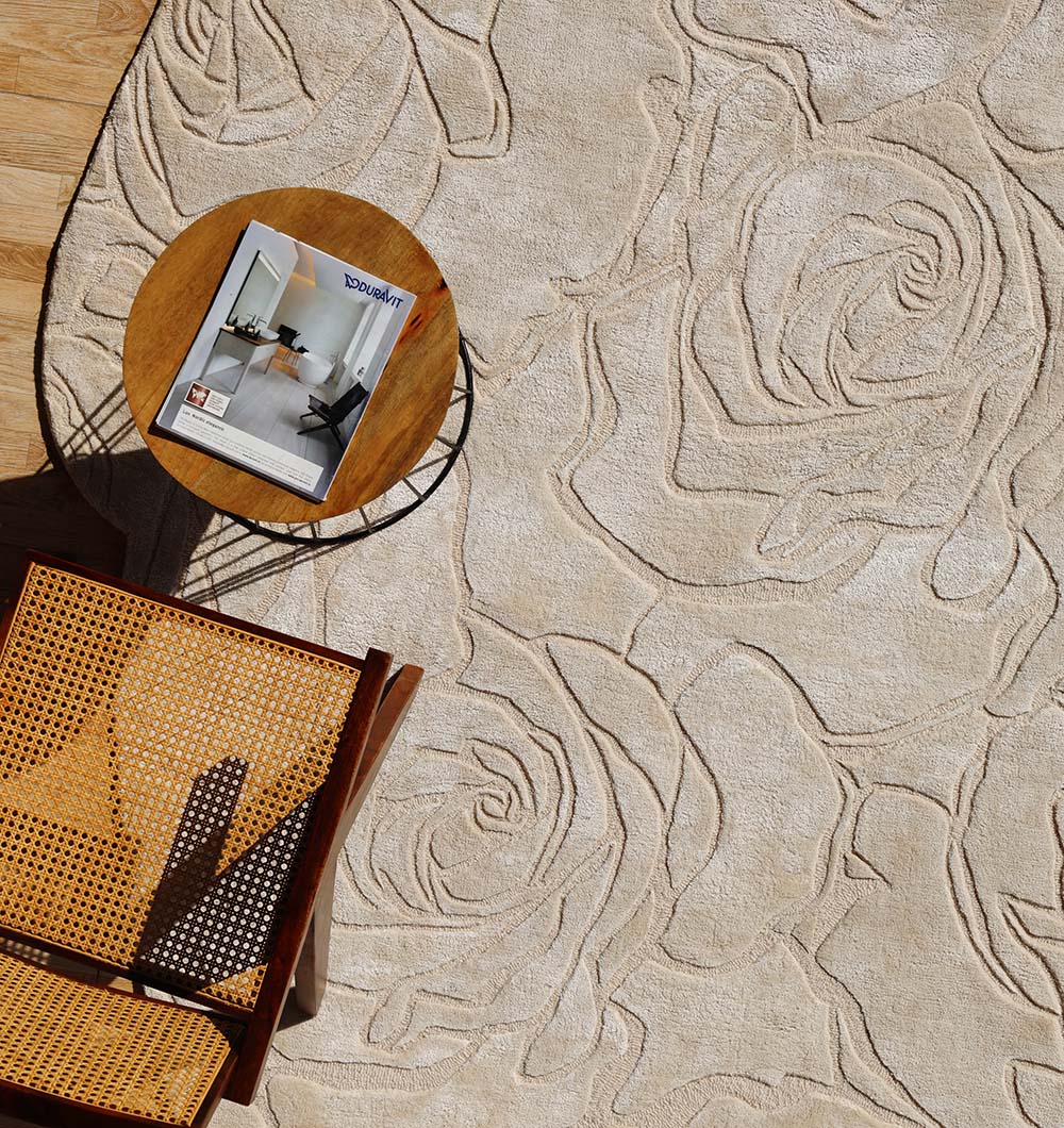 The RAMSHA HOME Home Decor Handmade Area Rug - HTWV-156, a light beige floral wool rug with a carved texture, lies beside a wooden chair and round side table. Sunlight highlights its elegant raised pattern and hand-tufted craftsmanship.