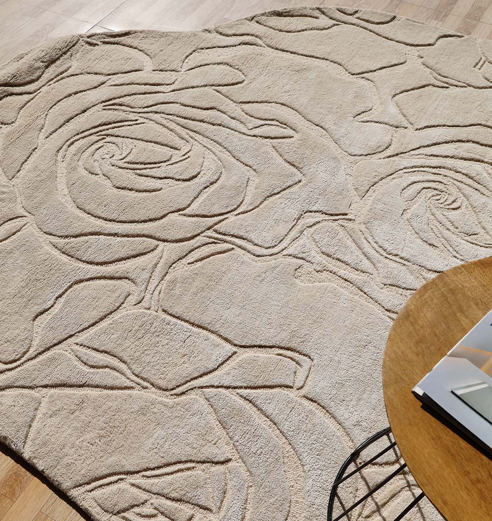 The RAMSHA HOME Home Decor Handmade Area Rug - HTWV-156, featuring a beige floral design with a raised texture and abstract rose patterns, lies on a light wooden floor beside a round table, softly illuminated by sunlight.