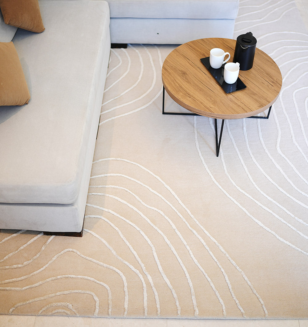 A round wooden coffee table with a black tray, teapot, and two cups sits on the RAMSHA HOME Handmade Home Decor Area Rug – HTWV-157, which features curved white lines, placed next to a light grey sofa with brown cushions.