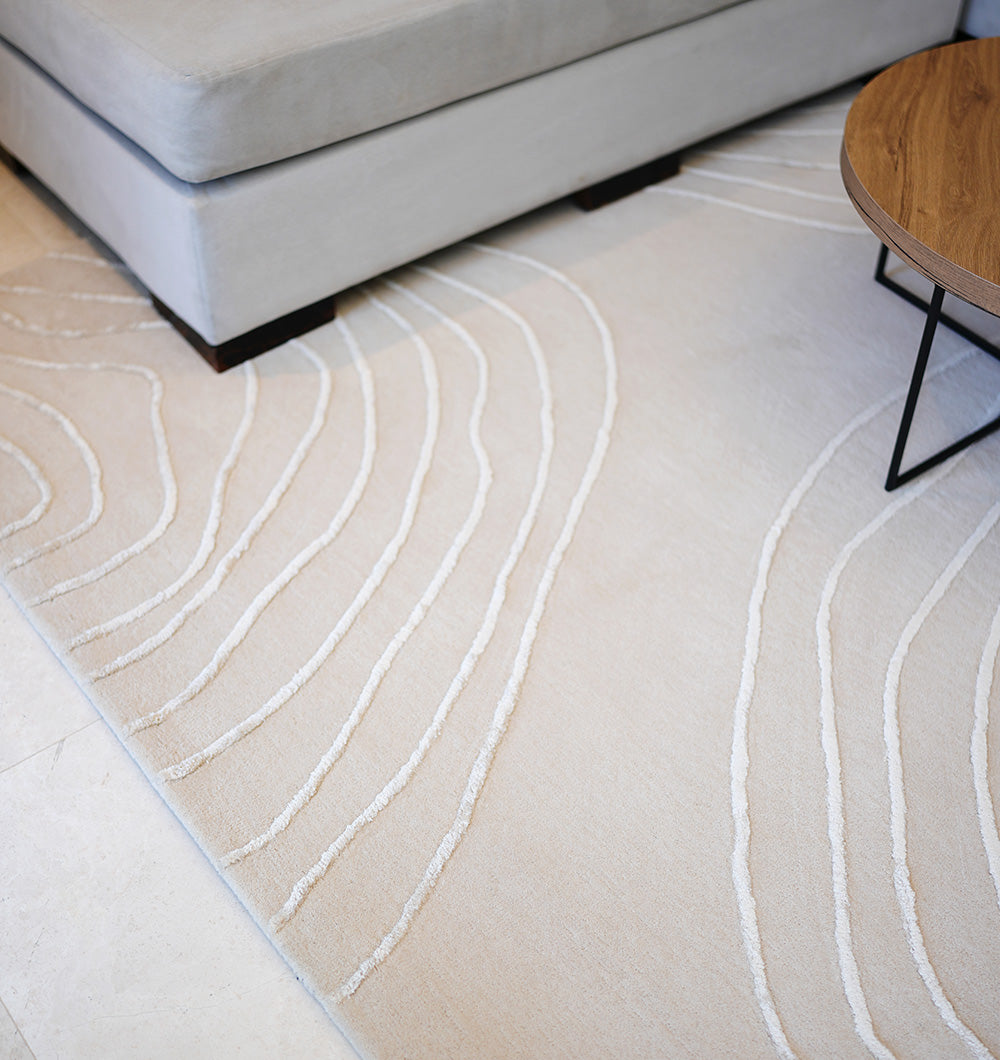 A close-up of the RAMSHA HOME Handmade Home Decor Area Rug – HTWV-157, featuring white curved lines, placed under a light gray sectional and a round wooden coffee table with black metal legs.