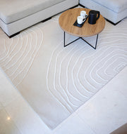 A modern living room highlights the RAMSHA HOME Handmade Home Decor Area Rug – HTWV-157 with abstract wavy lines, a round wooden coffee table with black metal legs, white cups, a black coffee pot, and a light gray sofa.