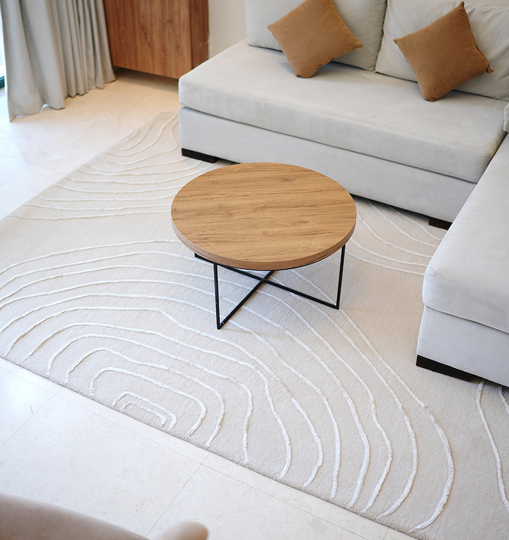 A modern living room featuring a pale beige sectional sofa, tan accent pillows, a round wooden coffee table with black metal legs, and the RAMSHA HOME Handmade Home Decor Area Rug – HTWV-157 in light tones with textured line patterns.