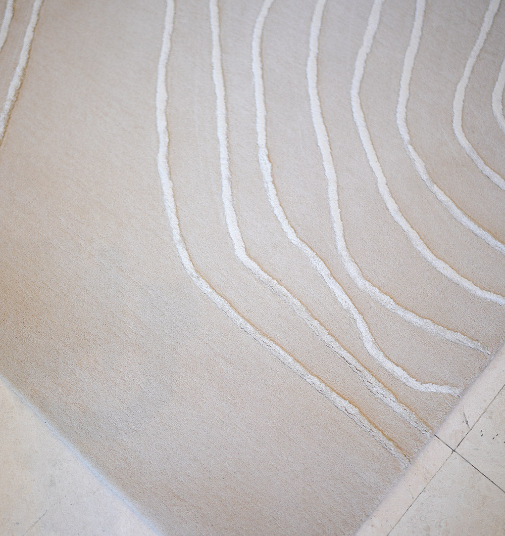 A close-up of the RAMSHA HOME Handmade Home Decor Area Rug – HTWV-157 in beige shows its soft, hand tufted texture with subtle, wavy raised lines forming an abstract pattern, set against a smooth, light-colored floor.