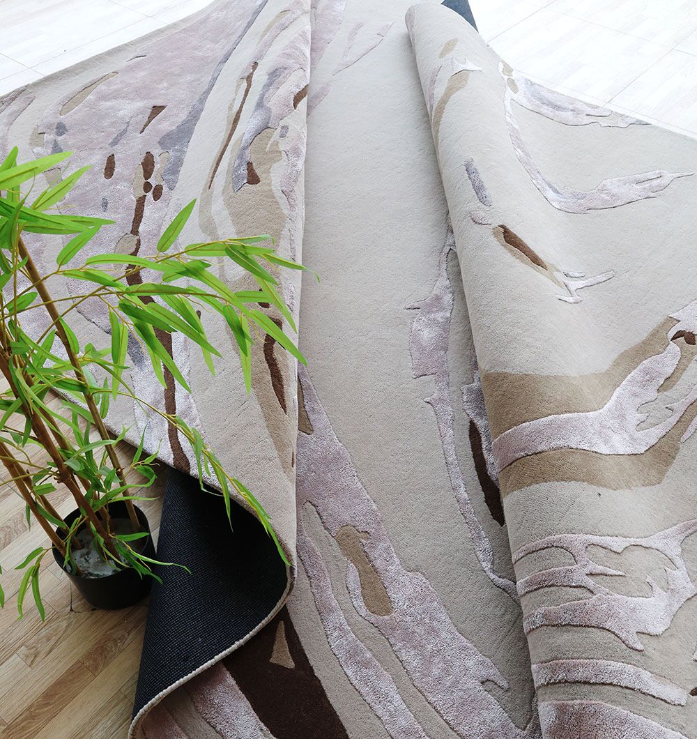 The RAMSHA HOME Multi Color Handmade Rug - HTWV 158 (300X400 cm) in beige, brown, and light purple lies folded on a light wooden floor next to a green potted bamboo plant.