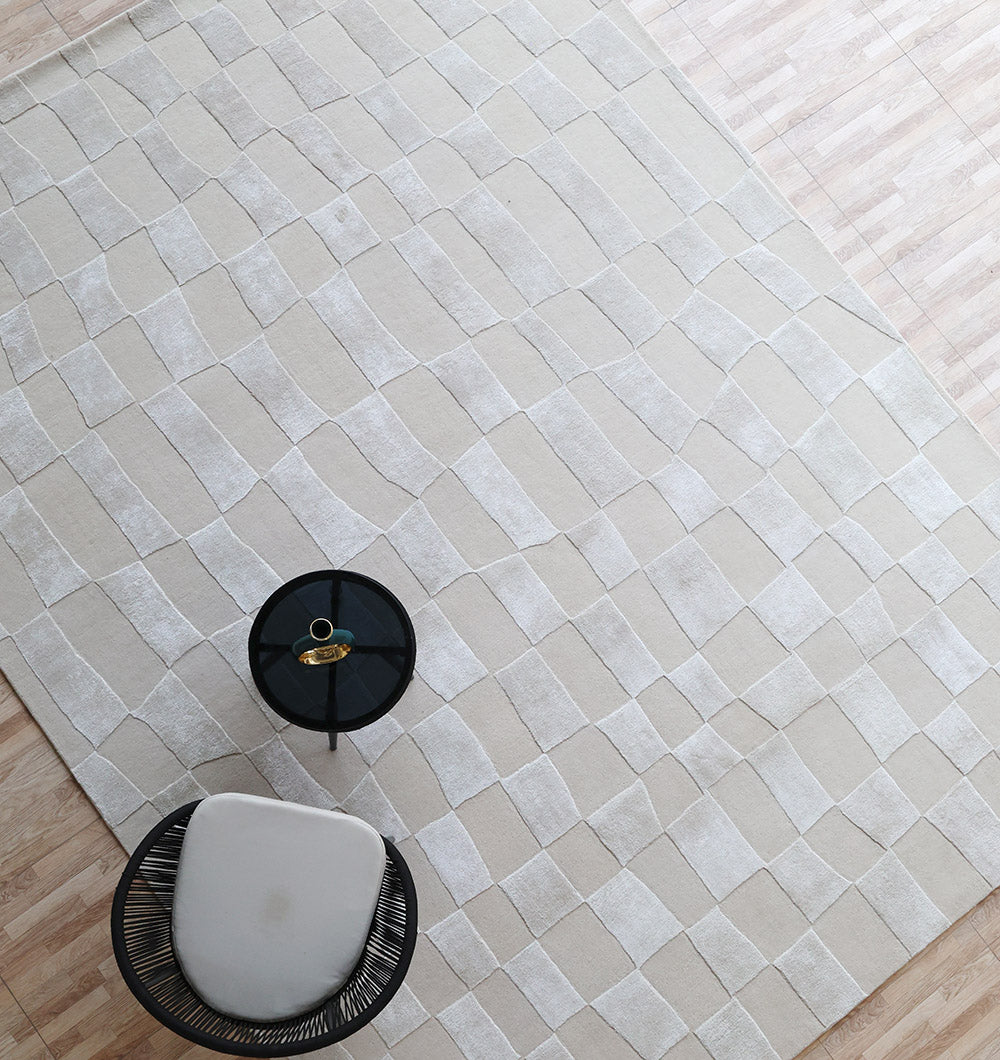 A top-down view of the RAMSHA HOME Light Beige Color Handmade Carpet – HTWV-159, featuring a modern design, placed on a wooden floor alongside a round black side table with decor and a contemporary curved chair with wire base.