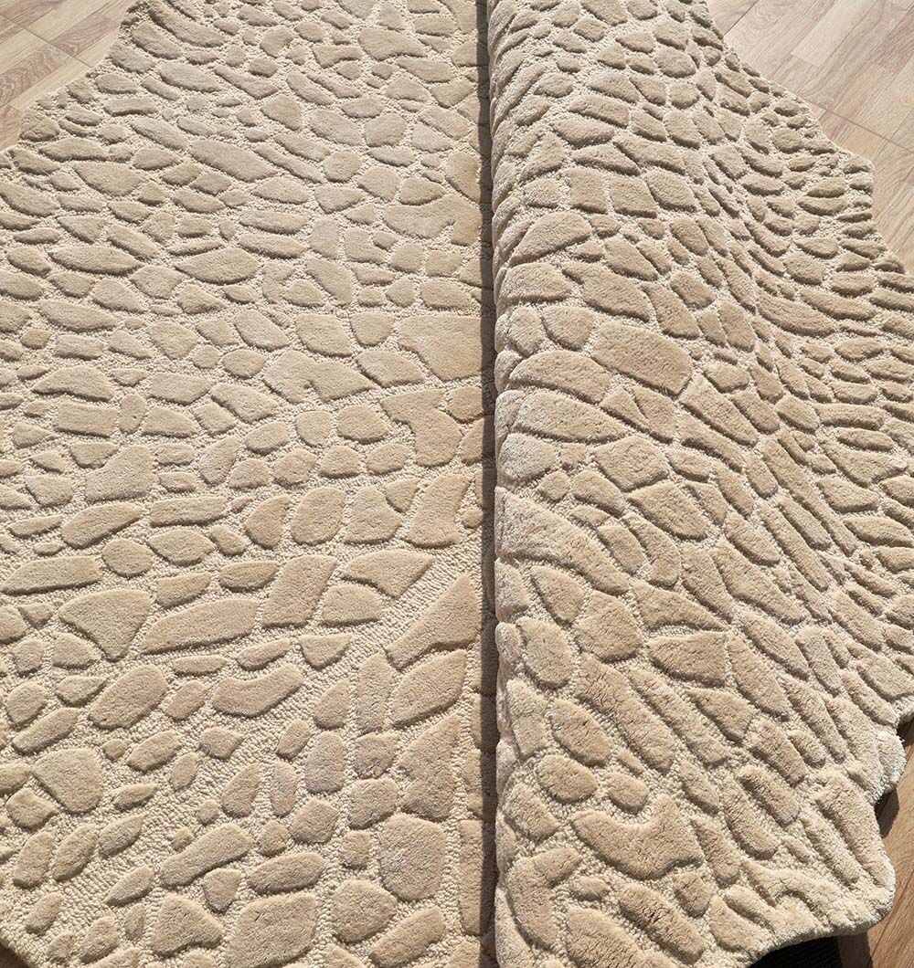 A close-up of the RAMSHA HOME Shaped Area Handmade Rug - HTWV 162 (200 cm), a beige hand-tufted wool rug with a textured, stone-inspired pattern, partially folded to display its thickness on a light wooden floor.
