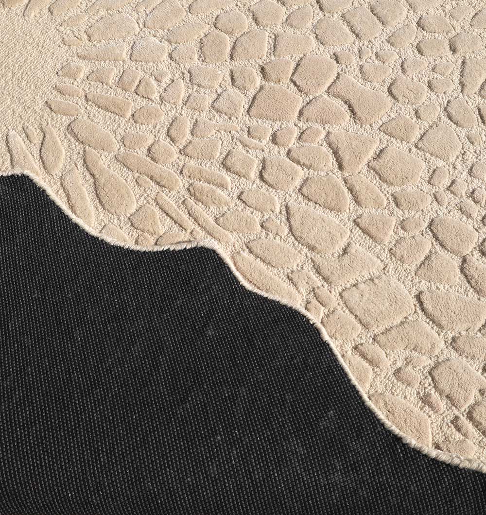 A close-up of the RAMSHA HOME Shaped Area Handmade Rug - HTWV 162 (200 cm) in beige shows its irregular, pebble-like textured wool pattern layered over a black fabric—ideal for those seeking a unique, hand-tufted rug.