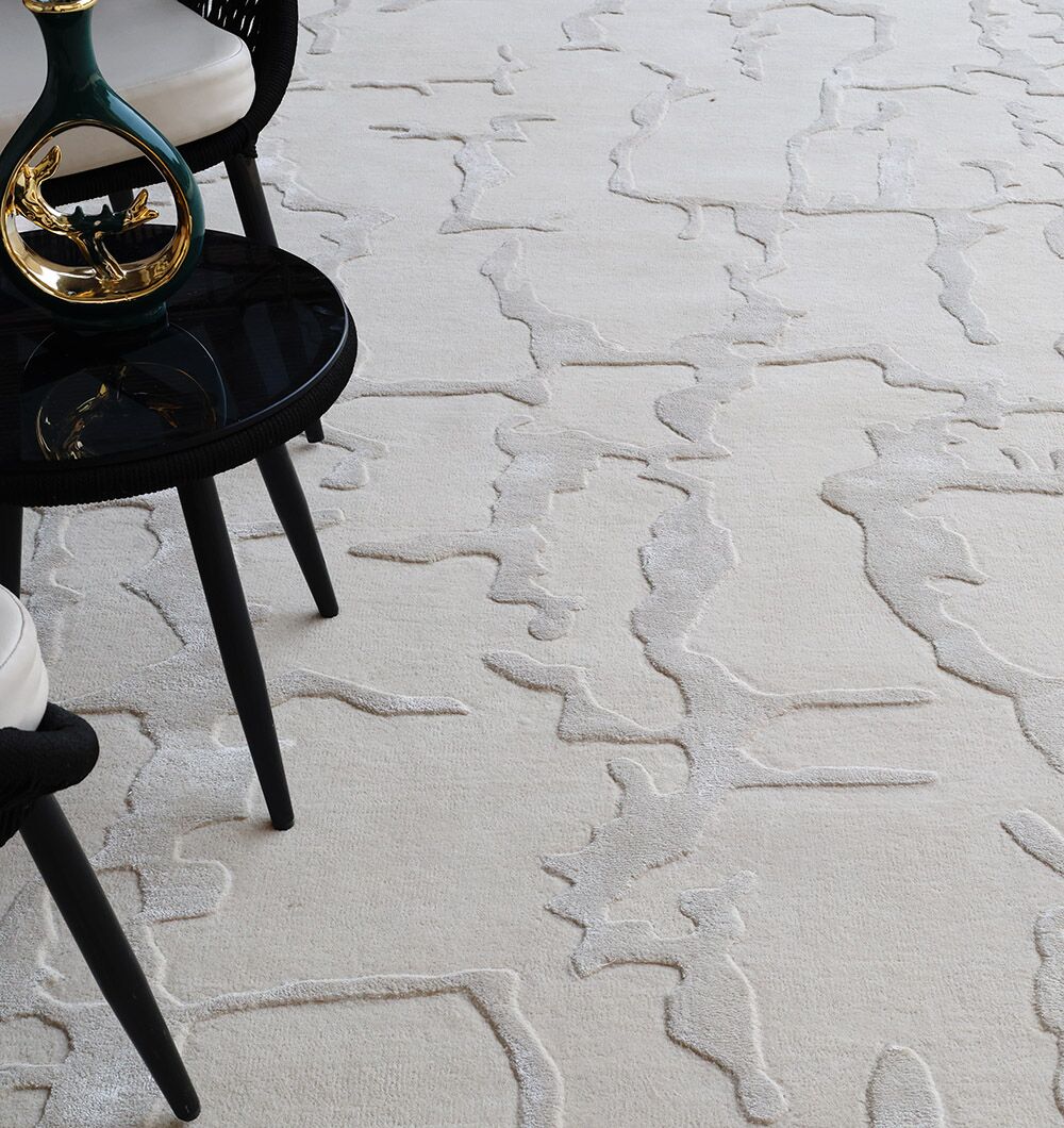 A close-up of the RAMSHA HOME Handmade Textured Crackle Pattern Leather Rug - HTWV 163 (300X400 cm) in beige, shown beside a black round side table with gold accents and part of a black chair with a white cushion.