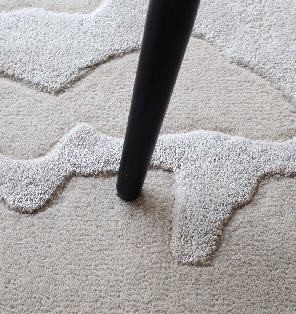 A close-up of a black furniture leg resting on the RAMSHA HOME Handmade Textured Crackle Pattern Leather Rug - HTWV 163 (300X400 cm), featuring beige wool viscose with irregular, raised white designs.