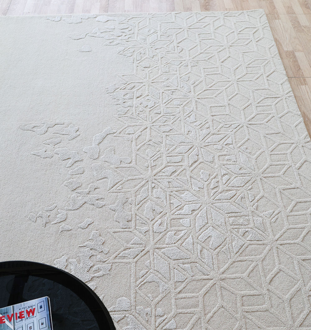 The RAMSHA HOME Handmade Beige Color Area Rug (HTWV 165, 200x300 cm) with a subtle geometric pattern lies on a light wood floor. A round black table holding magazines is placed in the lower left corner of the image.